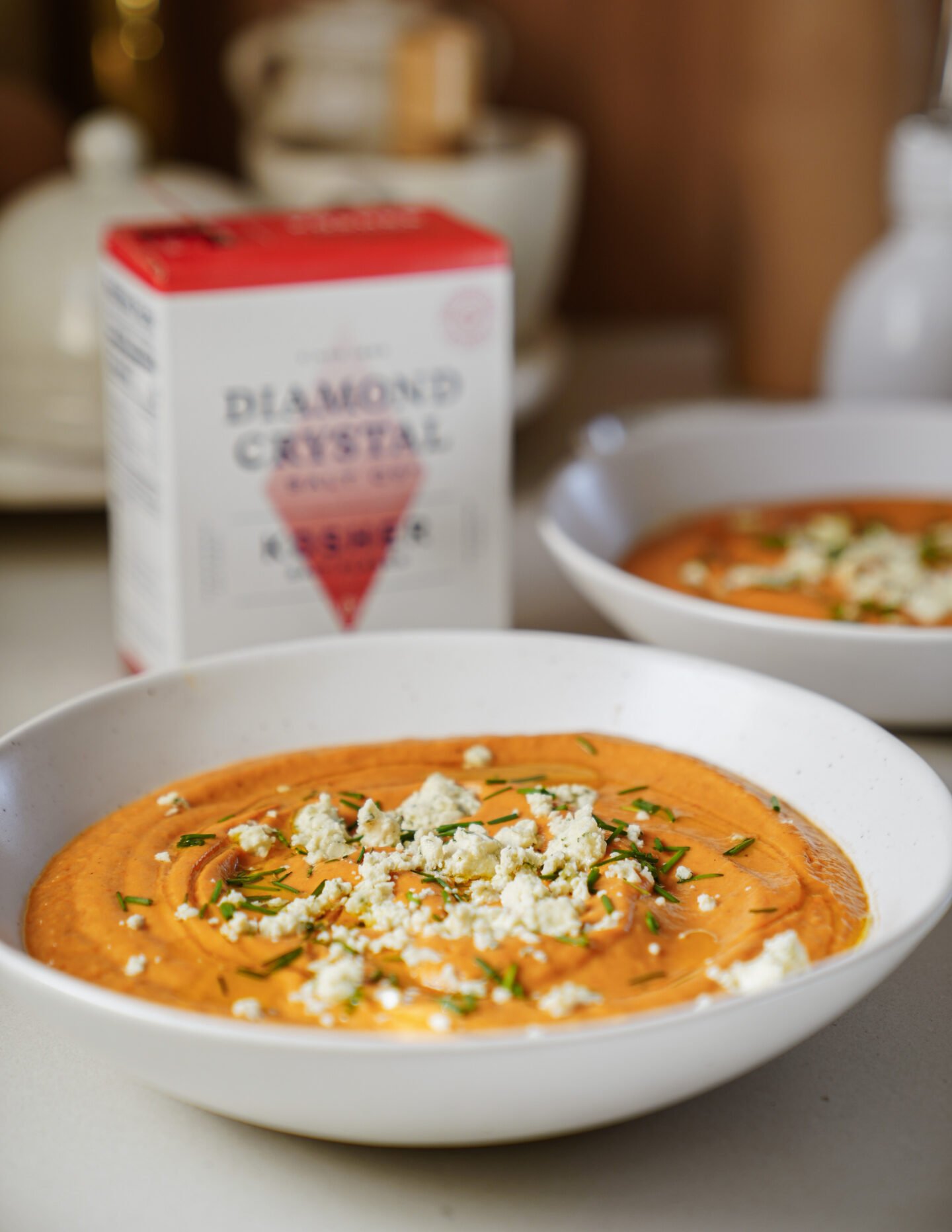 A bowl of creamy orange soup topped with crumbled cheese and chopped herbs sits on a table, with another similar bowl and a box of kosher salt visible in the background.