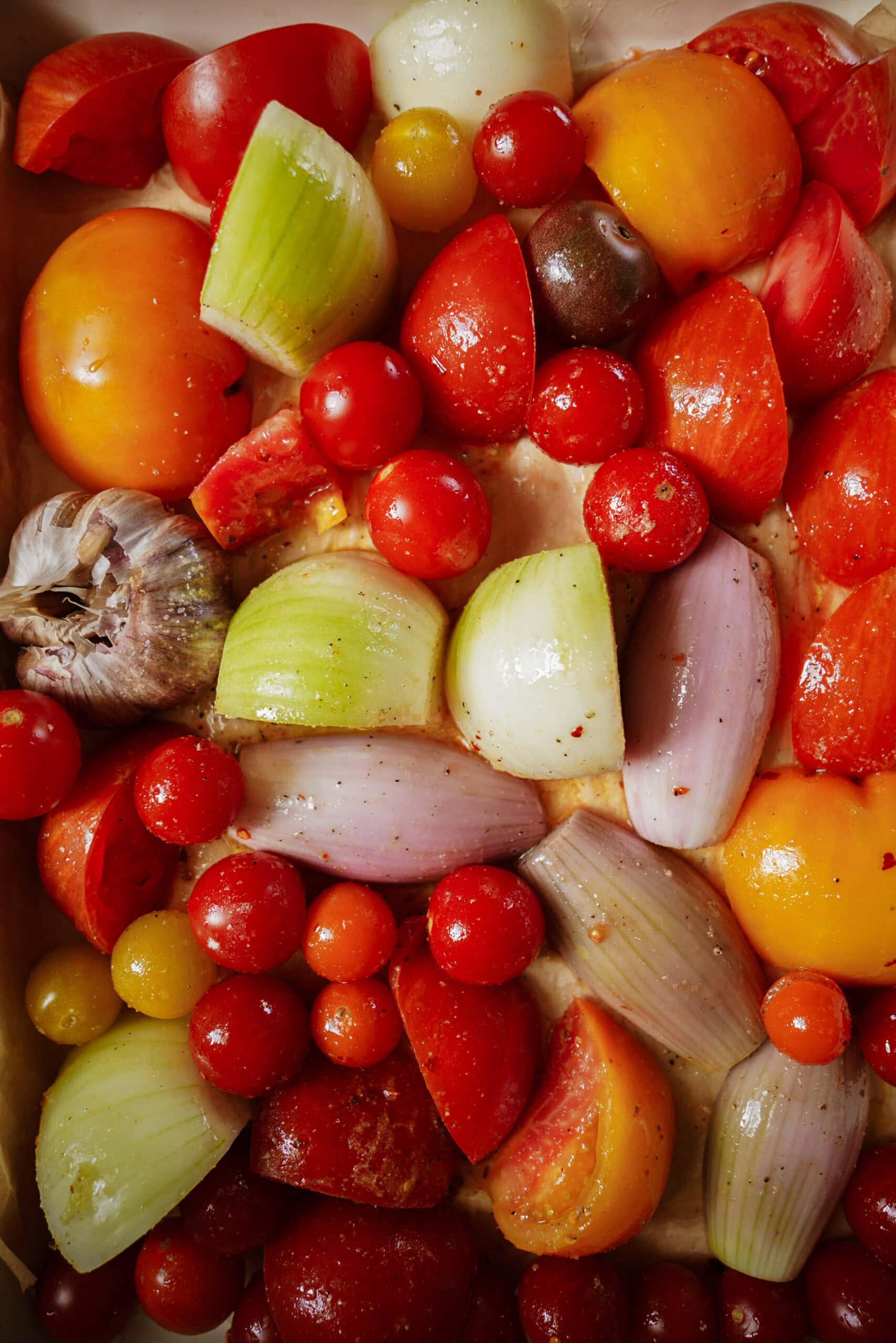 A colorful assortment of tomatoes, onions, shallots, and a head of garlic, all cut and arranged closely together on a baking tray for roasting.