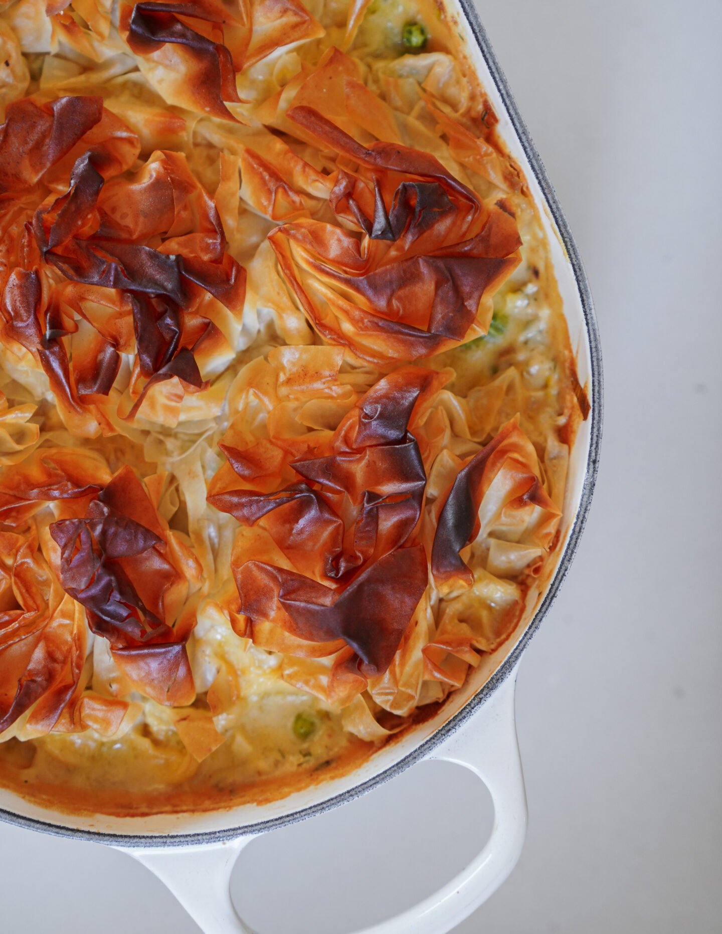 A close-up of a baked dish in a white casserole, topped with golden-brown, crinkled phyllo pastry. The pastry is crisp and layered, with some areas more browned than others.