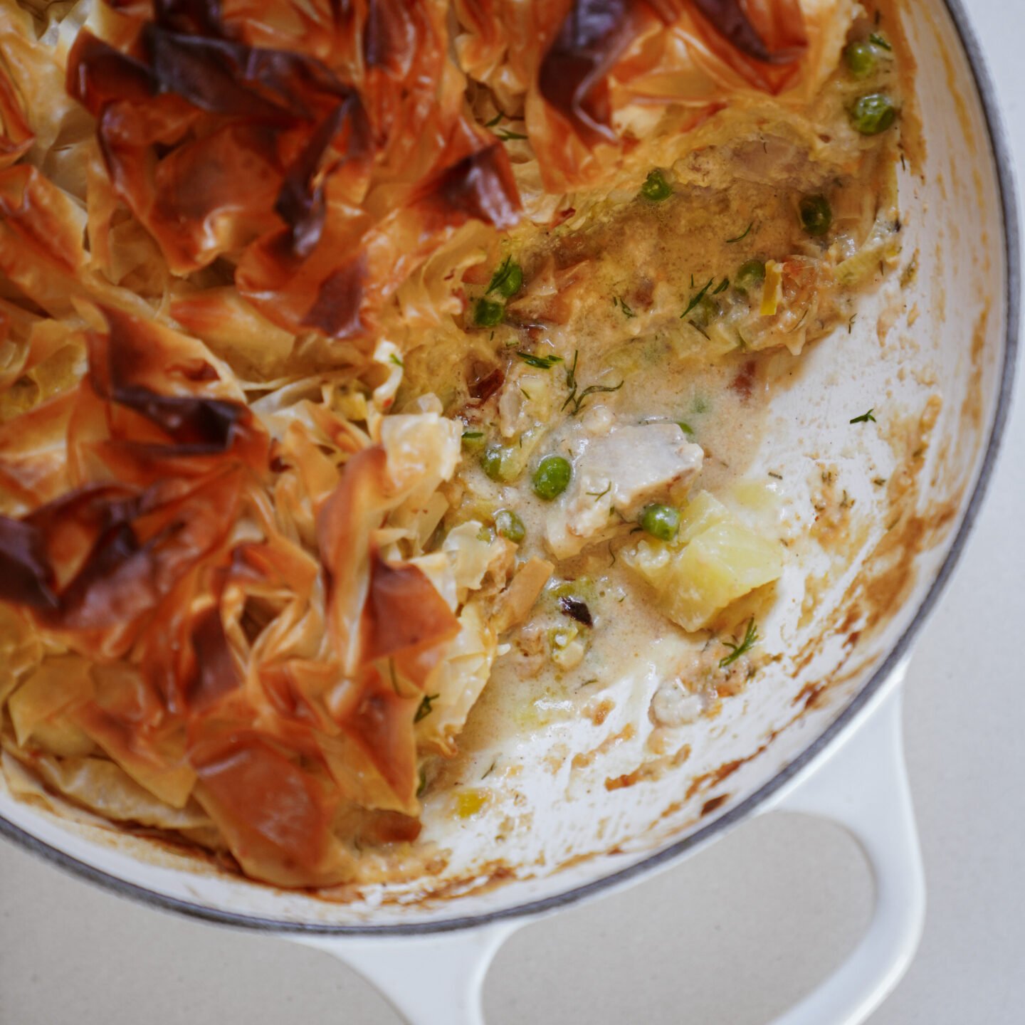 A white casserole dish containing a creamy pie with golden, crispy pastry on top. The filling includes chunks of meat, potatoes, peas, and herbs, with a portion removed, revealing the inside.