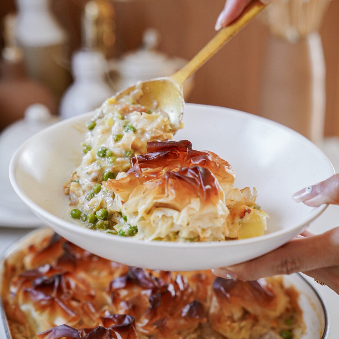 A hand serves a portion of golden-brown filo pastry-topped casserole with peas and vegetables onto a white plate using a gold spoon. The casserole dish is visible in the background.