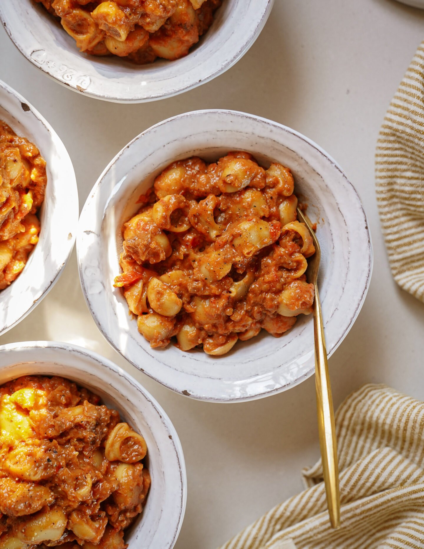 Four white bowls filled with shell pasta in a creamy tomato sauce are arranged on a light surface. One bowl has a gold spoon, and a beige-striped cloth is nearby. The food is warmly lit from above.