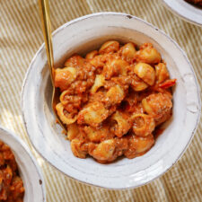 A bowl of creamy pasta with shell-shaped noodles, ground meat, and a rich tomato sauce, served with a gold-colored fork on a striped beige surface.