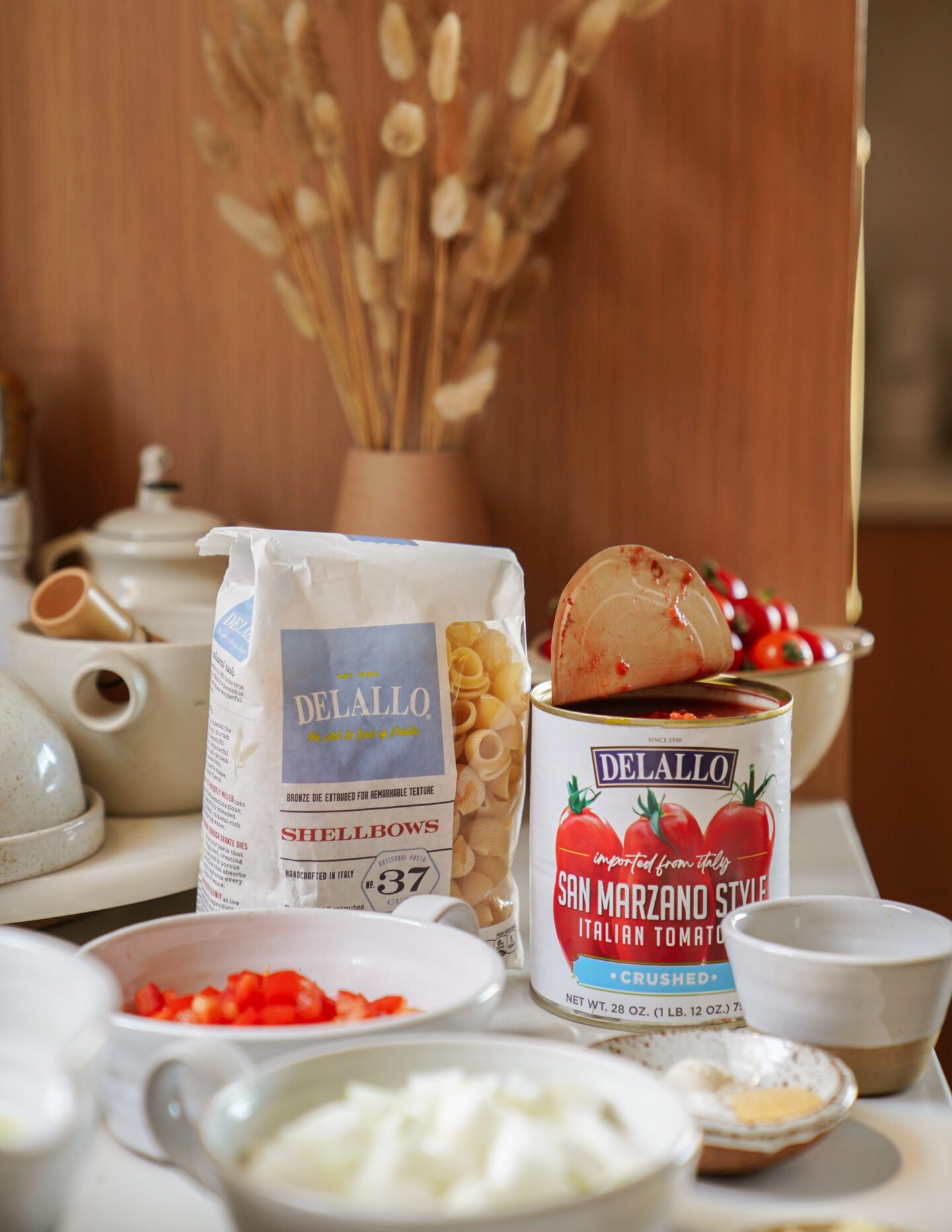 A kitchen counter with pasta, an open can of San Marzano tomatoes, bowls of chopped onions and tomatoes, and cooking utensils, with a bag of shellbows pasta and dried flowers in the background.