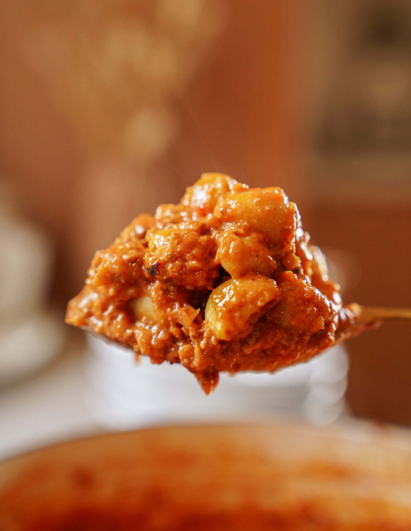 A close-up of a spoonful of chunky tomato-based stew or pasta sauce, featuring pieces of meat and gnocchi, held above a pot with a blurred background.