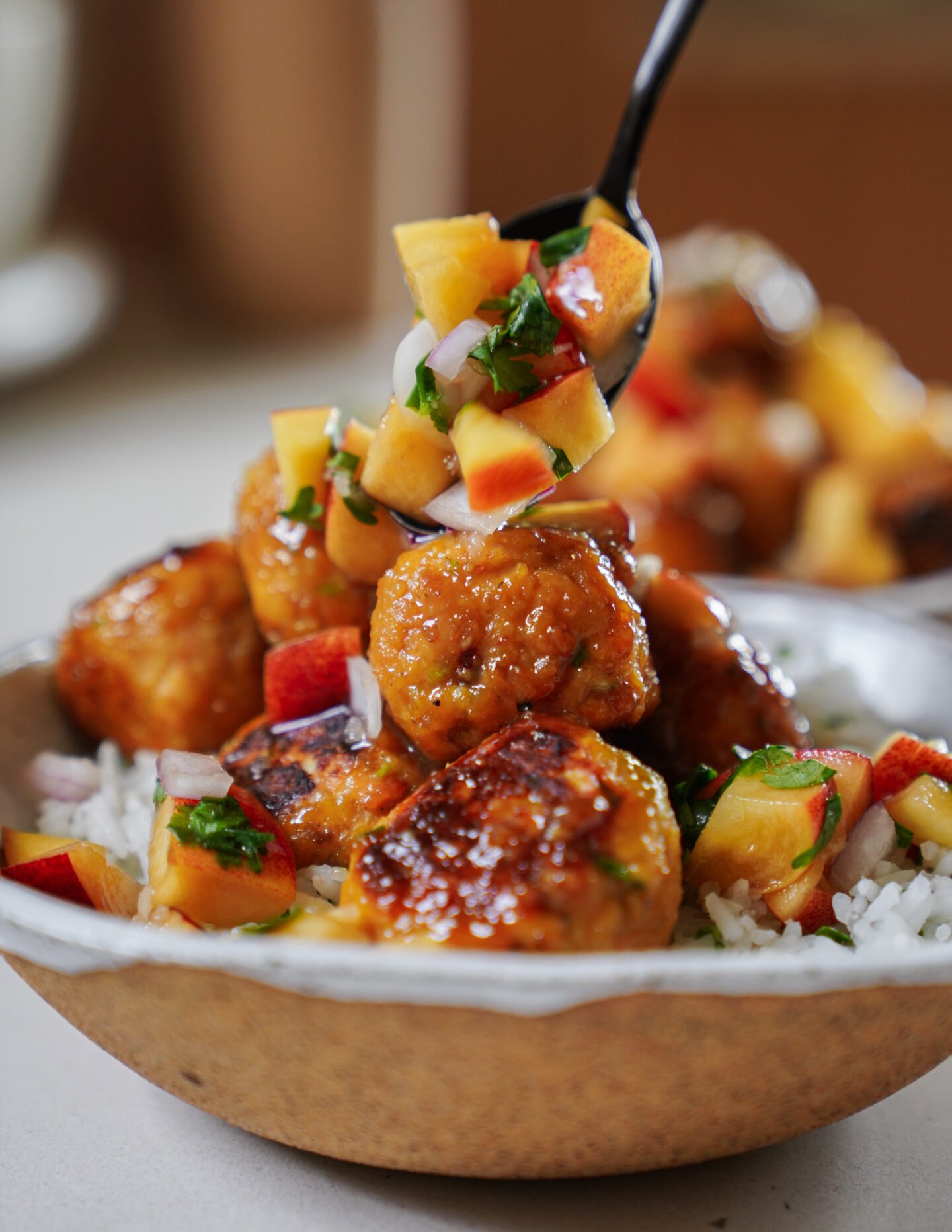 A bowl of glazed meatballs served over white rice, topped with a colorful fresh peach salsa. A spoon is adding more salsa to the meatballs, highlighting vibrant pieces of peach, red onion, and cilantro.