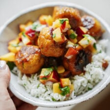 A hand holds a bowl of white rice topped with browned meatballs and a colorful peach salsa made of diced peaches, red onion, and fresh herbs.