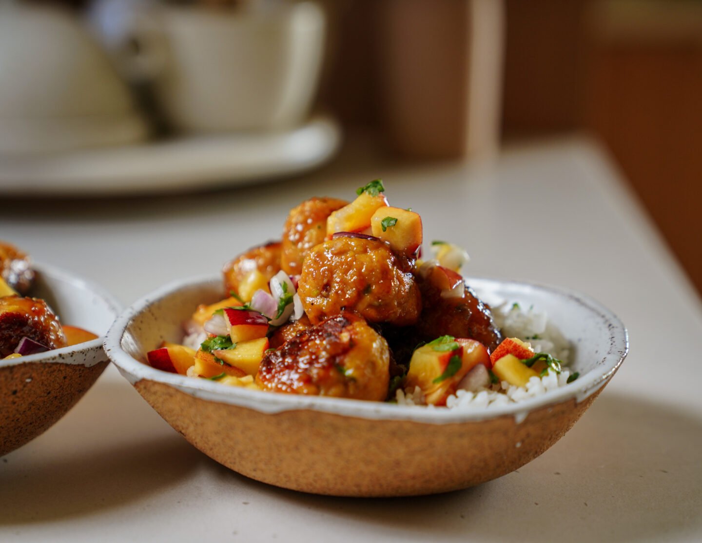 A bowl of white rice topped with glazed meatballs and a colorful fruit salsa, including diced peaches and herbs, served in a rustic ceramic dish on a light countertop.