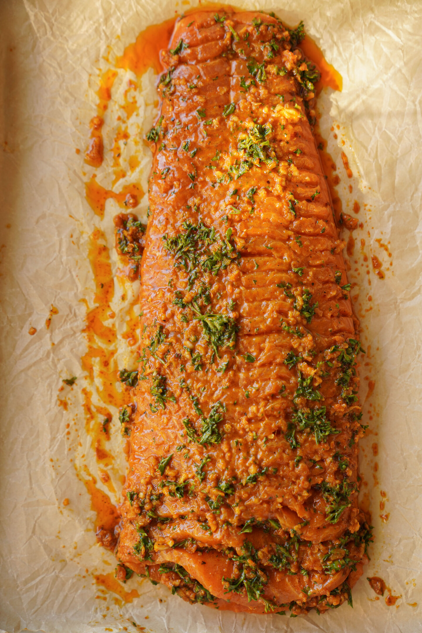 A large, seasoned fillet of salmon covered in an orange marinade with chopped herbs, placed on parchment paper in a baking tray.
