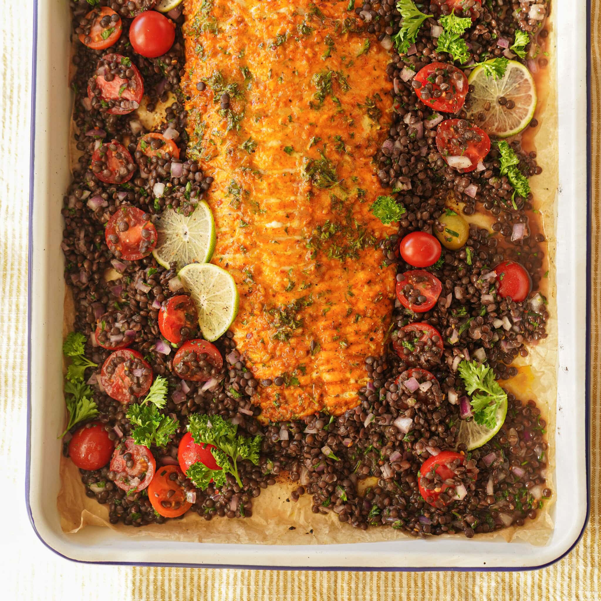 A baked salmon fillet garnished with herbs is surrounded by black lentils, cherry tomatoes, parsley, and lime slices in a large rectangular baking dish. The dish sits on a light yellow striped surface.