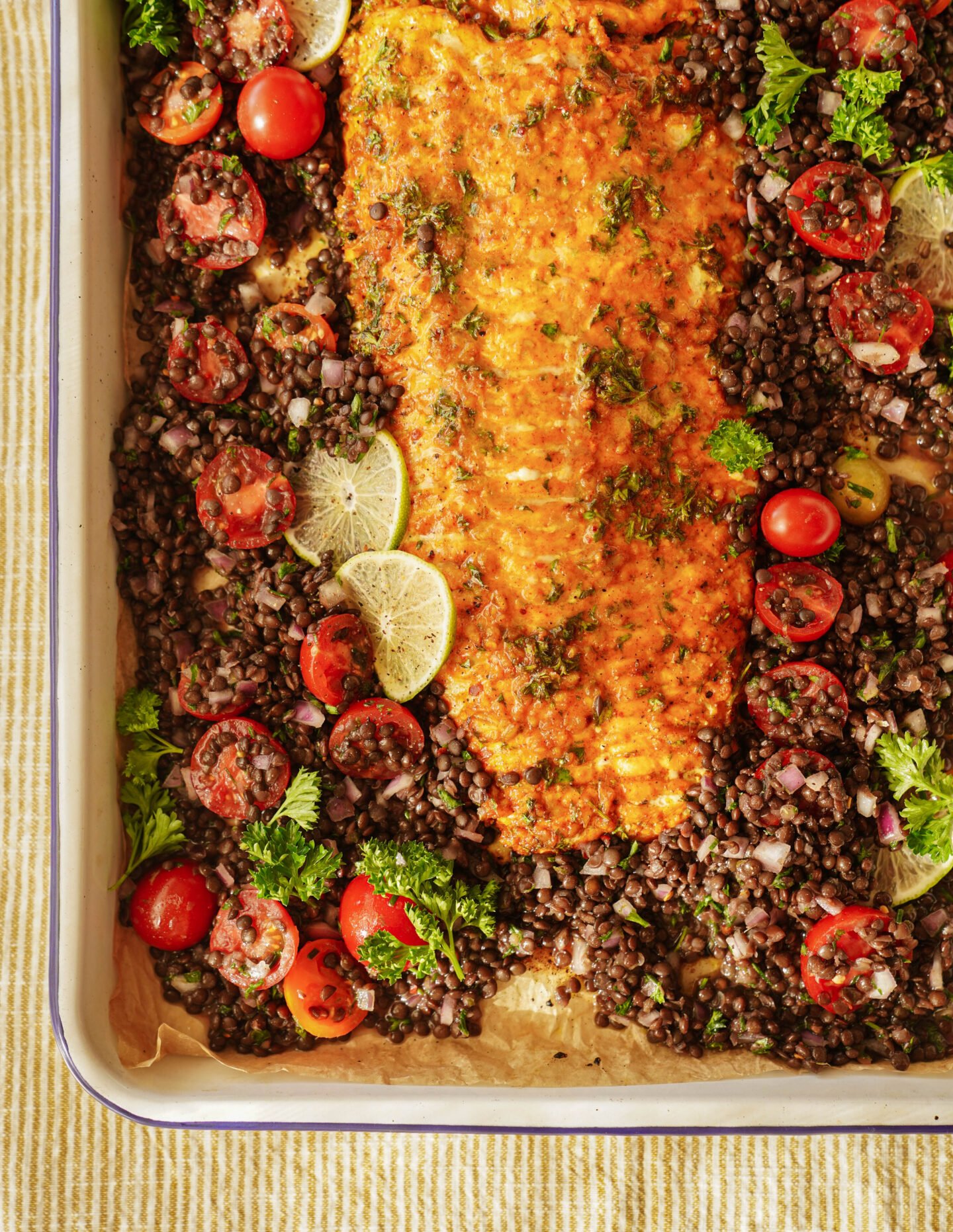 A baked salmon fillet topped with herbs is surrounded by a mixture of black lentils, cherry tomatoes, red onion, parsley, and lime slices on a parchment-lined baking sheet.