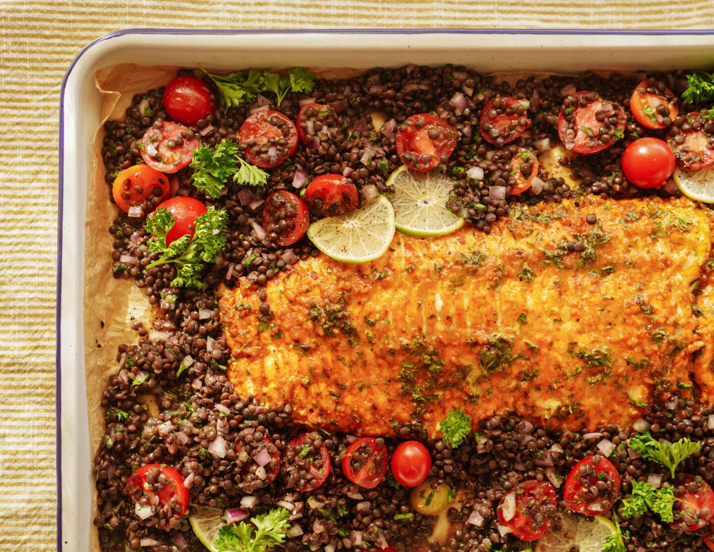 A baked salmon fillet topped with herbs is surrounded by a mixture of black lentils, cherry tomatoes, red onion, parsley, and lime slices on a parchment-lined baking sheet.
