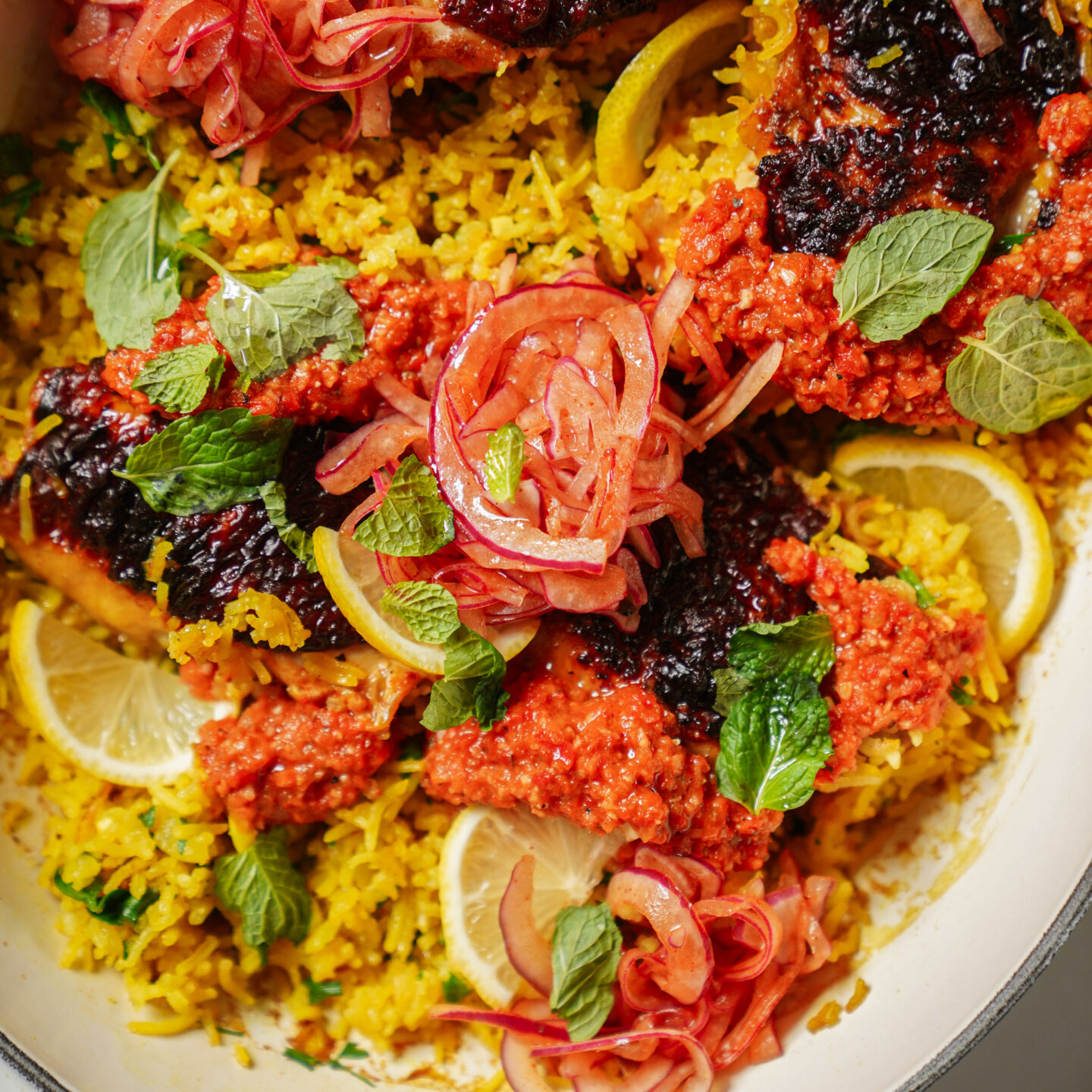 A close-up of a dish with yellow rice, topped with pieces of fish covered in red and dark sauces, garnished with pickled onions, fresh mint leaves, and lemon slices.