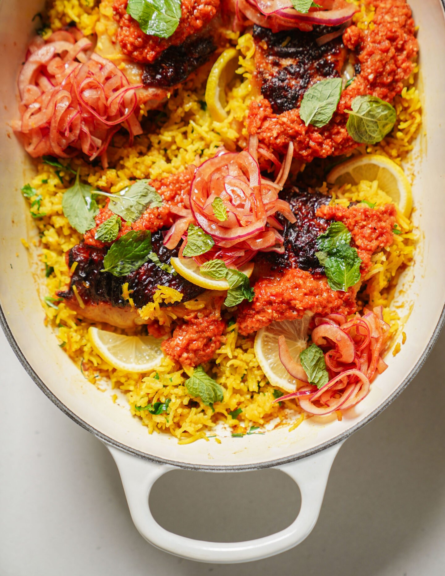 A white dish filled with yellow rice, garnished with lemon slices, grilled fish topped with red sauce, pickled red onions, and fresh mint leaves.