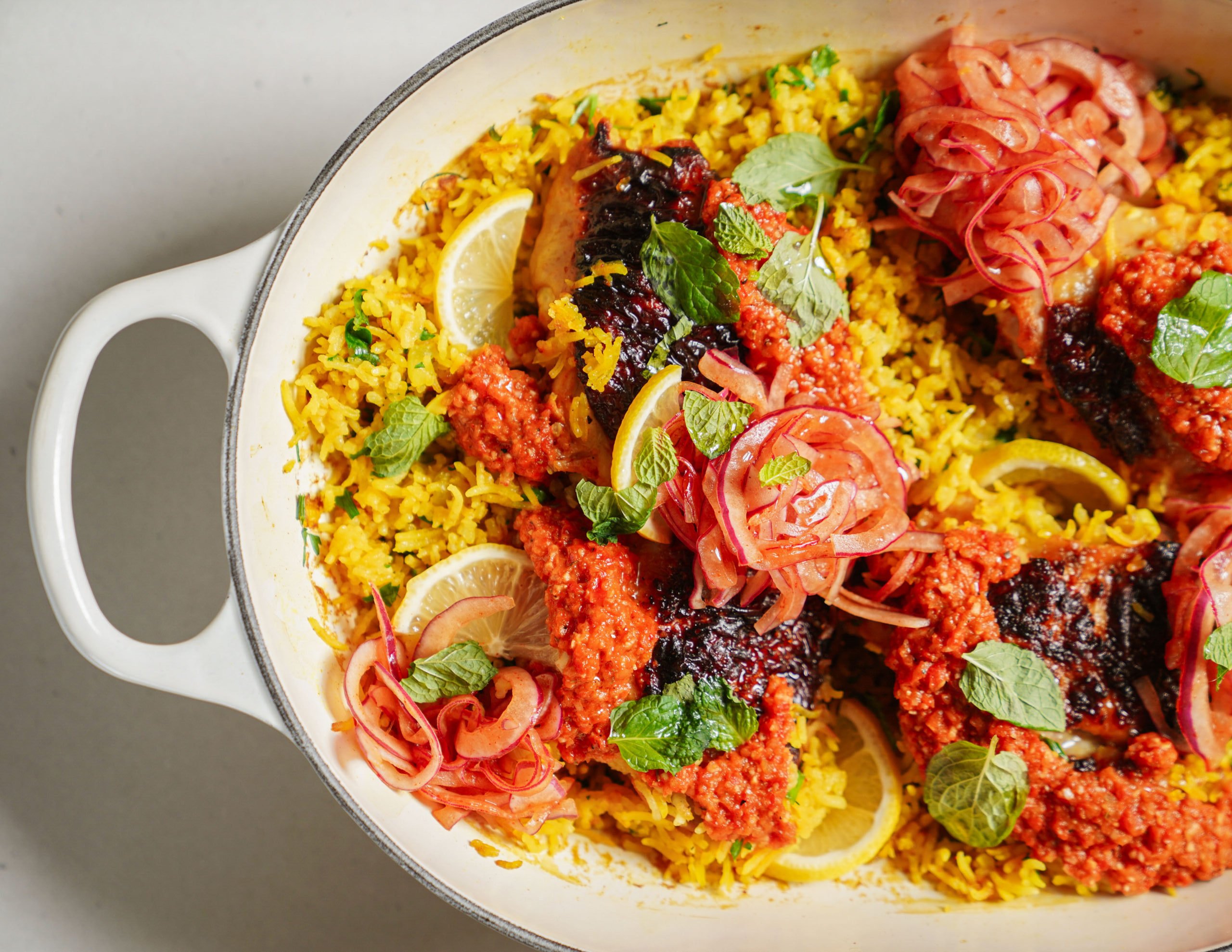 A white dish filled with yellow rice, garnished with lemon slices, grilled fish topped with red sauce, pickled red onions, and fresh mint leaves.