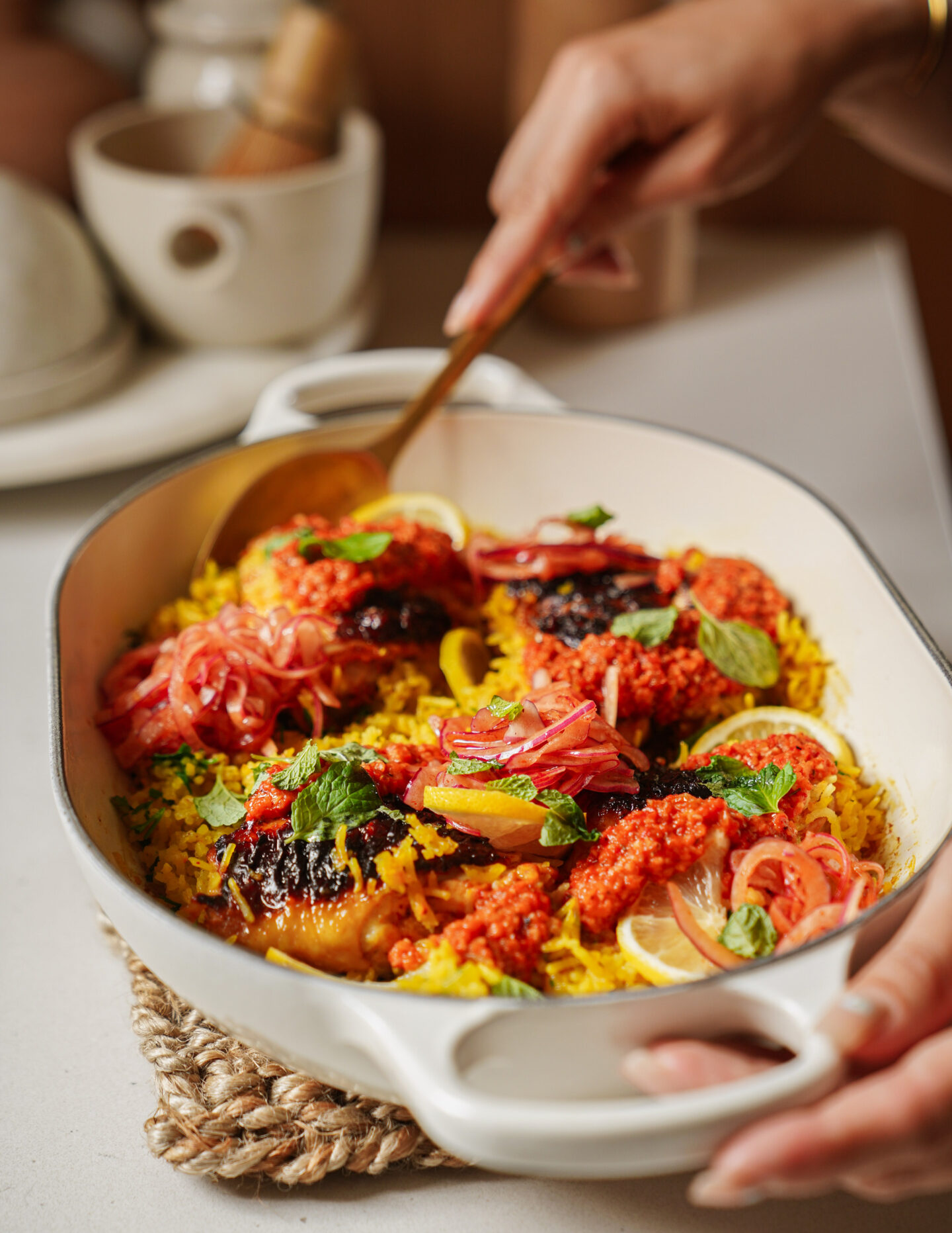 A hand serves yellow rice and baked chicken topped with red sauce, pickled onions, lemon slices, and fresh herbs from a white casserole dish. The dish sits on a woven trivet on a kitchen counter.