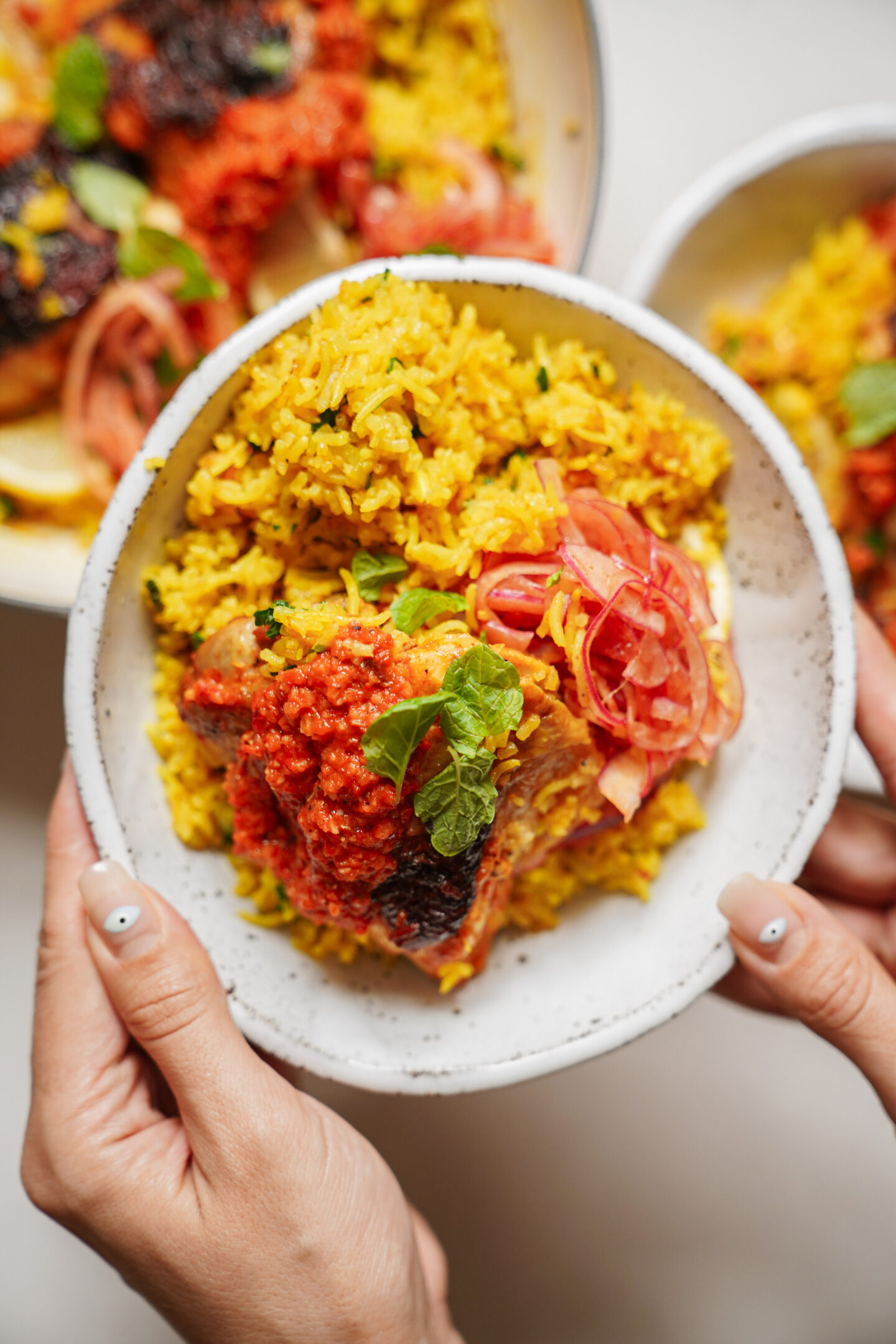 A person holds a bowl of yellow rice topped with grilled meat, red sauce, pickled onions, and a mint leaf, with more bowls of similar food in the background.