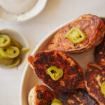 A close-up of several browned bread slices on a plate, each topped with sliced pickled jalapeños. A small bowl of extra jalapeño slices sits nearby on a light surface.