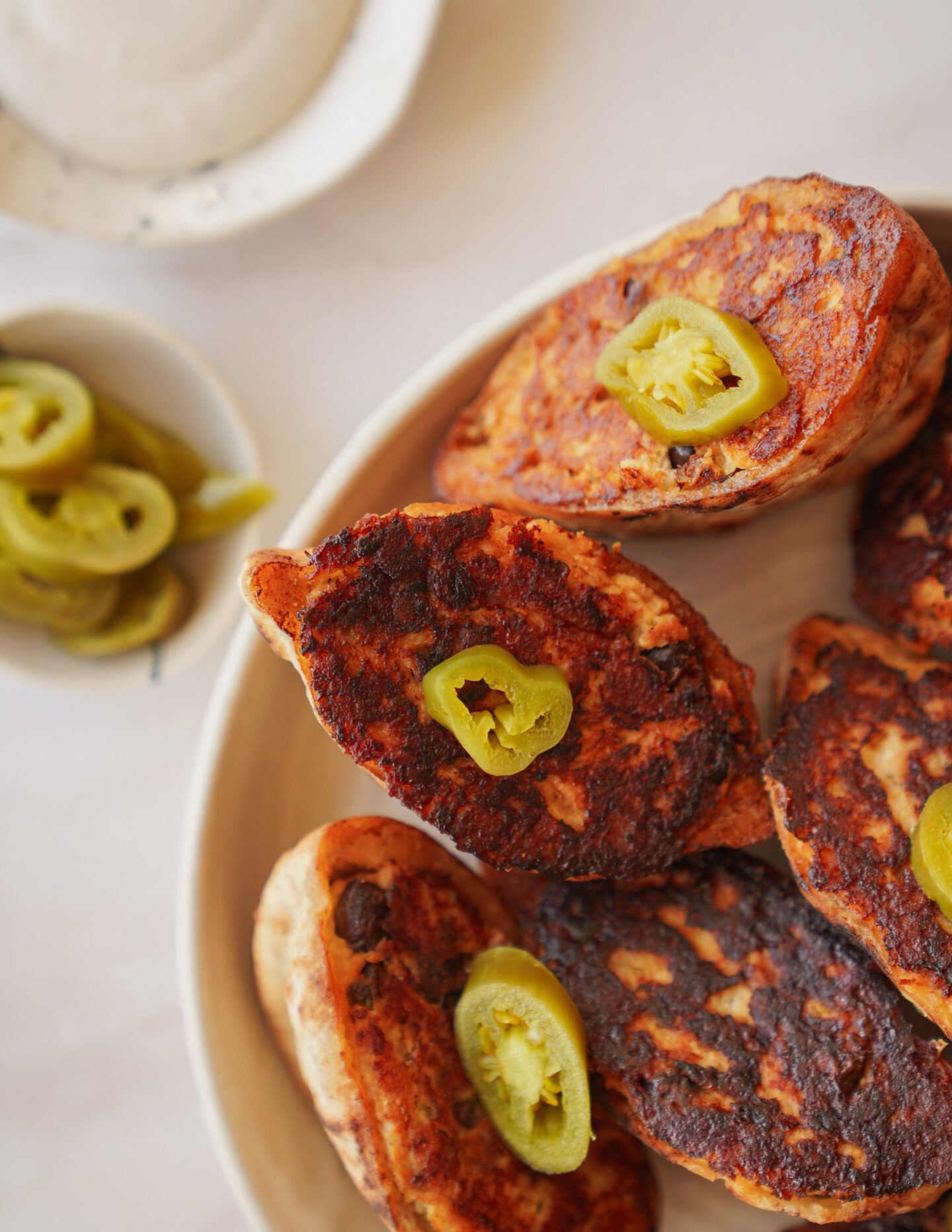 A plate of browned, oval-shaped fritters or patties, each topped with a slice of jalapeño pepper. A small bowl with extra jalapeño slices is nearby on a light-colored surface.