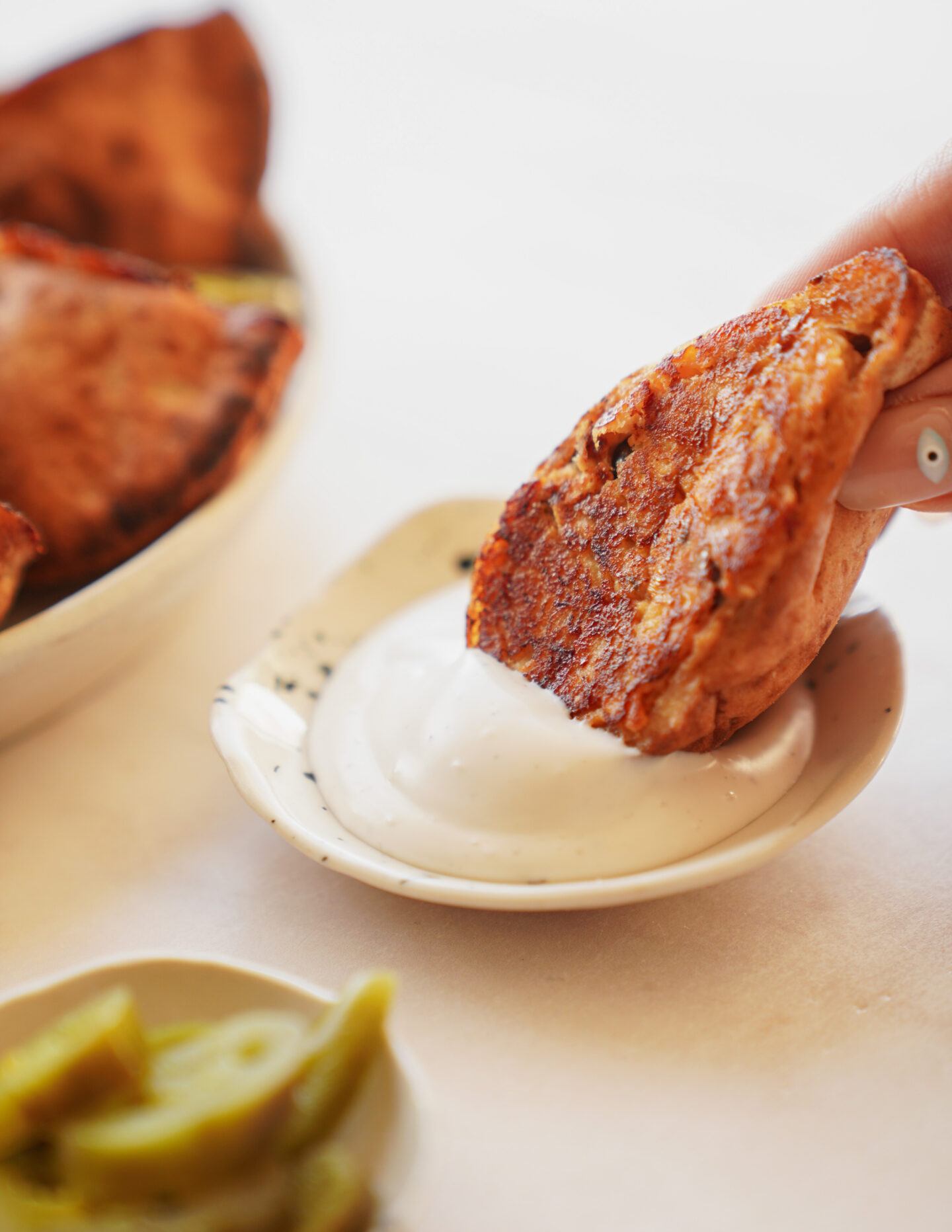 A hand dips a crispy, golden-brown food item into a small dish of creamy white sauce. A bowl of similar items and sliced green pickles are visible nearby.