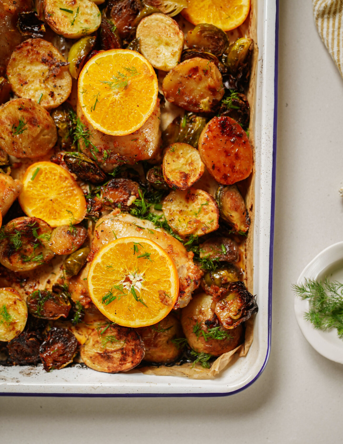 A baking dish filled with roasted potatoes, Brussels sprouts, and chicken pieces, topped with orange slices and sprinkled with fresh herbs. A small plate with dill is visible on the side.