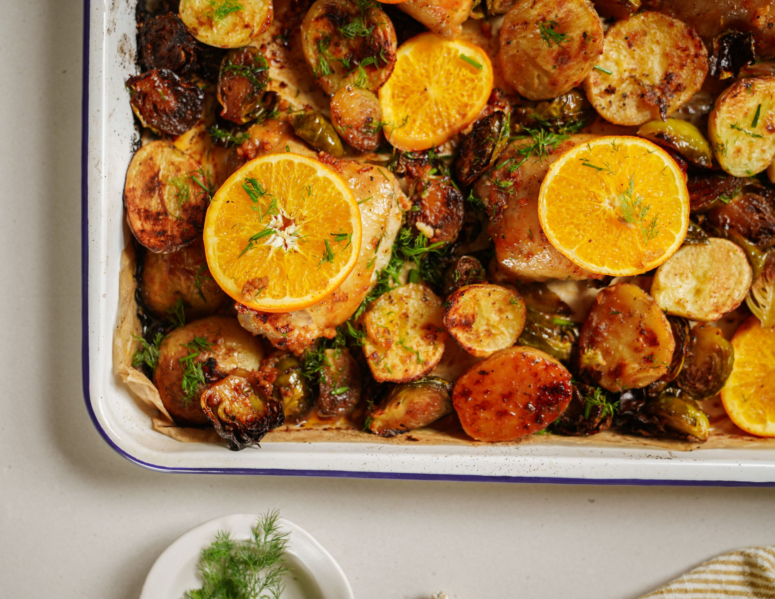 A baking dish filled with roasted potatoes, Brussels sprouts, and chicken pieces, topped with orange slices and sprinkled with fresh herbs. A small plate with dill is visible on the side.