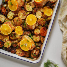 A baking tray filled with roasted chicken, sliced potatoes, Brussels sprouts, and garnished with orange and lemon slices, set on a light surface next to a yellow-striped cloth and a small bowl of fresh herbs.