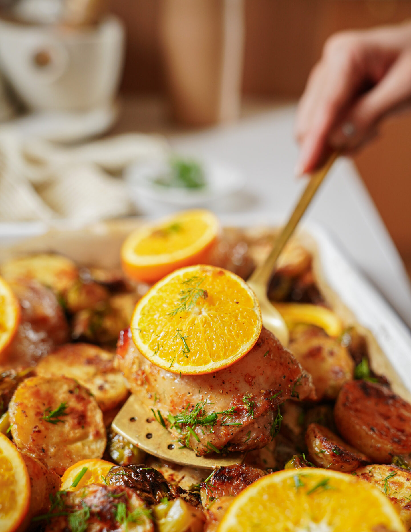 A hand uses a spatula to serve roasted chicken with golden potatoes, garnished with fresh dill and orange slices, from a baking dish. The scene looks appetizing and vibrant.