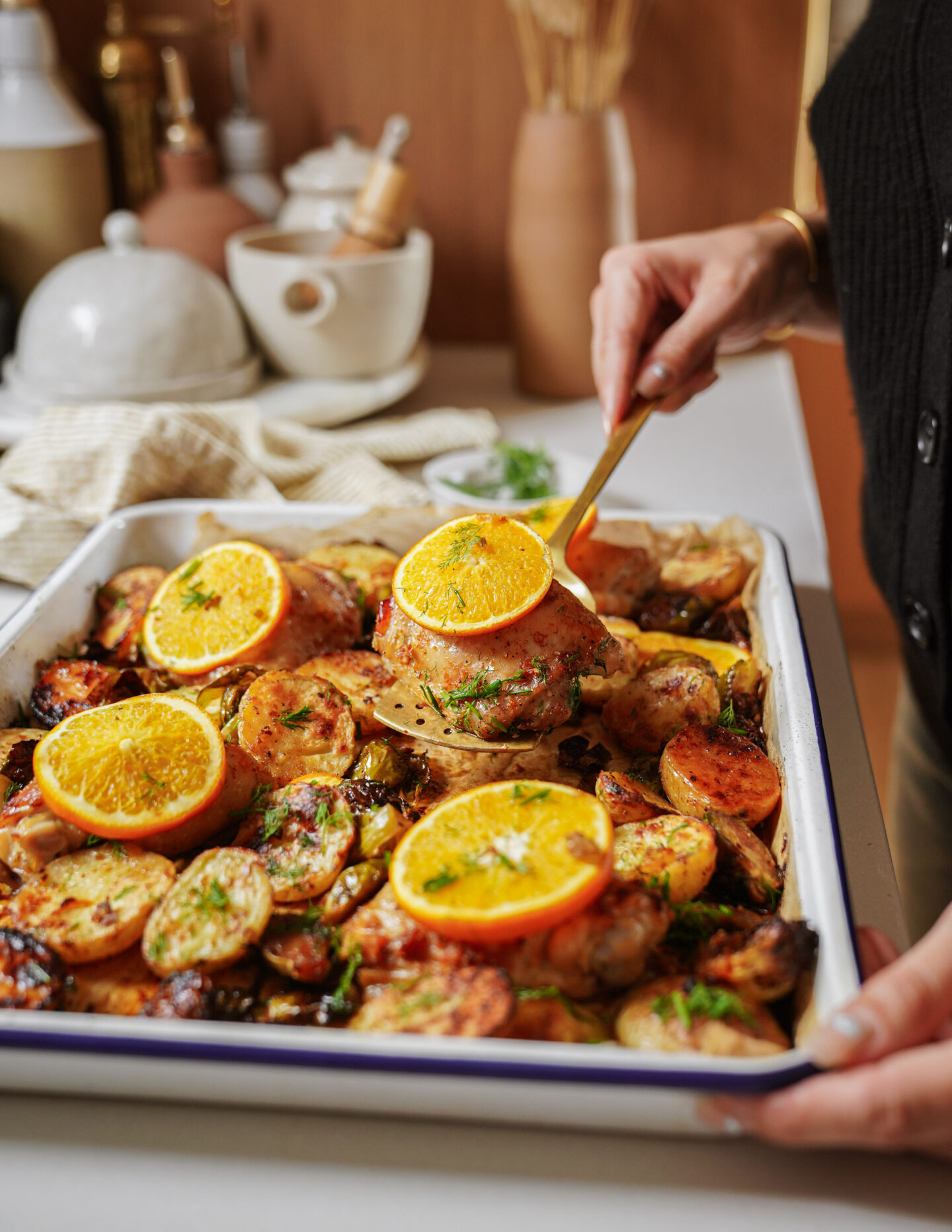 A person serves roasted chicken and potatoes from a baking dish, garnished with fresh herbs and orange slices, in a cozy kitchen setting.