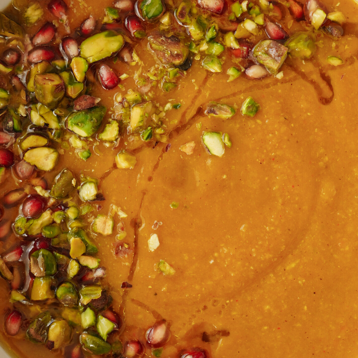 A close-up of a bowl of creamy soup garnished with chopped pistachios, pomegranate seeds, and a drizzle of oil. The vibrant toppings contrast with the smooth, orange-brown soup base.