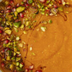 A close-up of a bowl of creamy soup garnished with chopped pistachios, pomegranate seeds, and a drizzle of oil. The vibrant toppings contrast with the smooth, orange-brown soup base.
