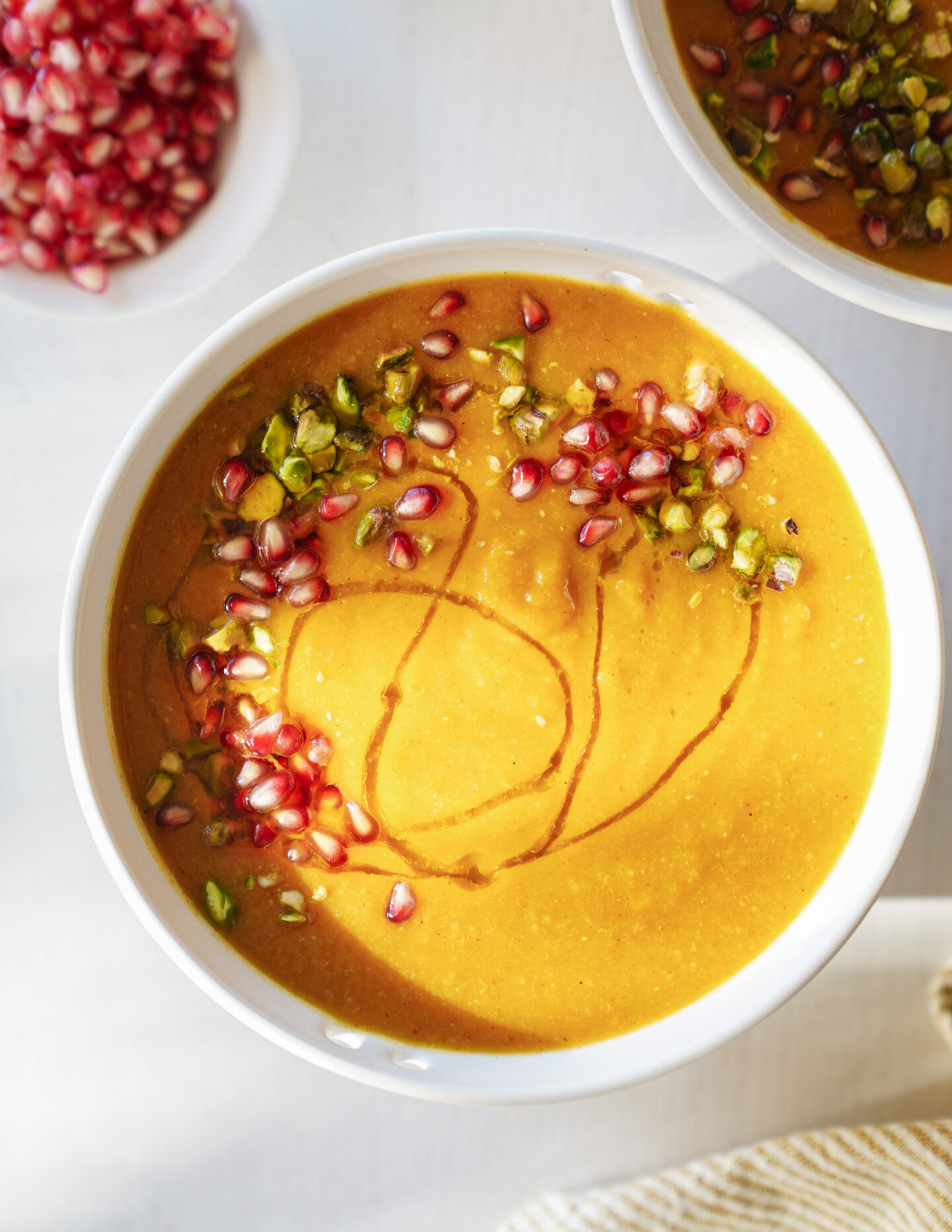 A bowl of creamy orange soup garnished with pomegranate seeds, chopped green pistachios, and a drizzle of oil. A small bowl of pomegranate seeds is visible in the background.