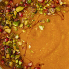 A close-up of a bowl of creamy soup garnished with chopped pistachios, pomegranate seeds, and a drizzle of oil. The vibrant toppings contrast with the smooth, orange-brown soup base.