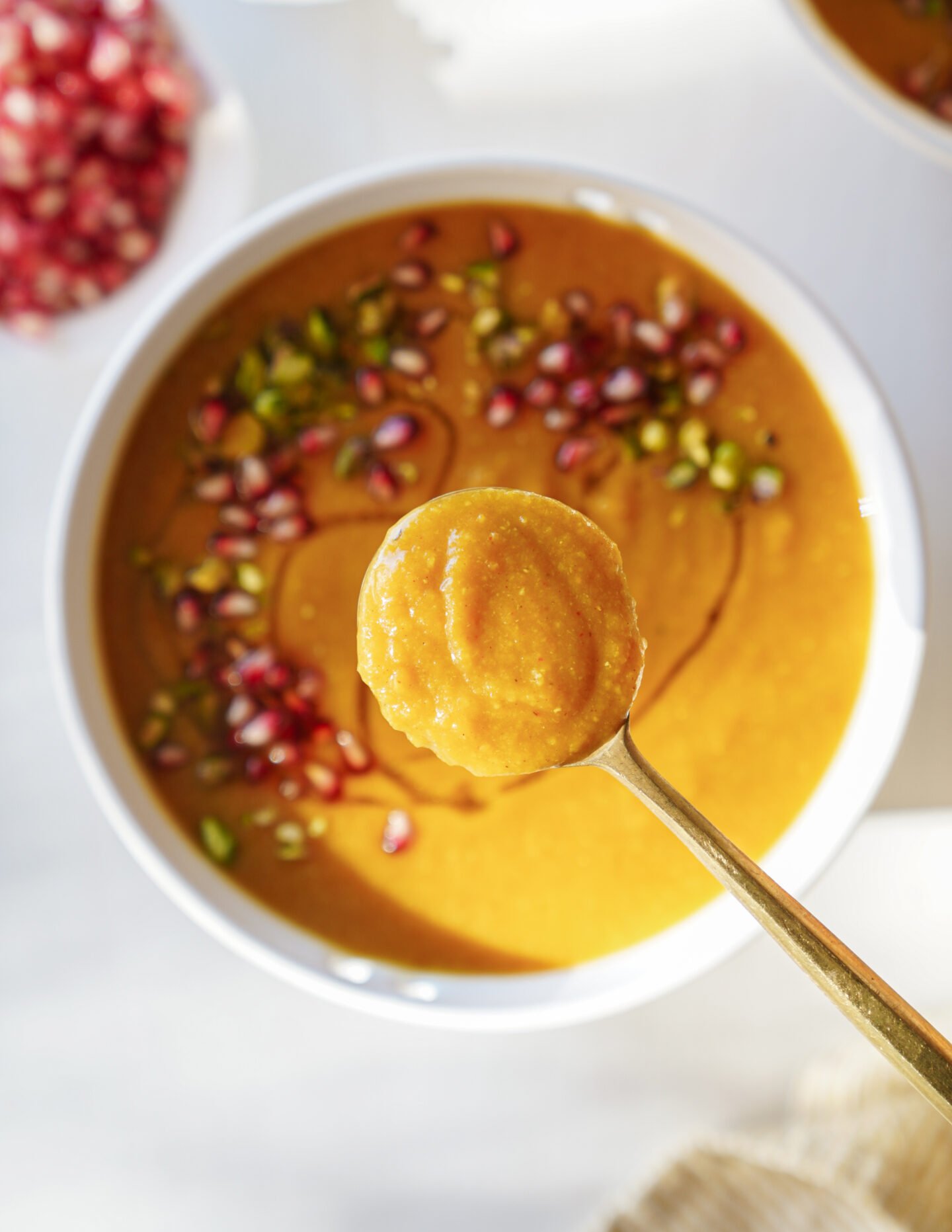 A close-up of a spoonful of orange soup held above a bowl, garnished with pomegranate seeds, green herbs, and a drizzle of oil. The bowl is set on a white surface.