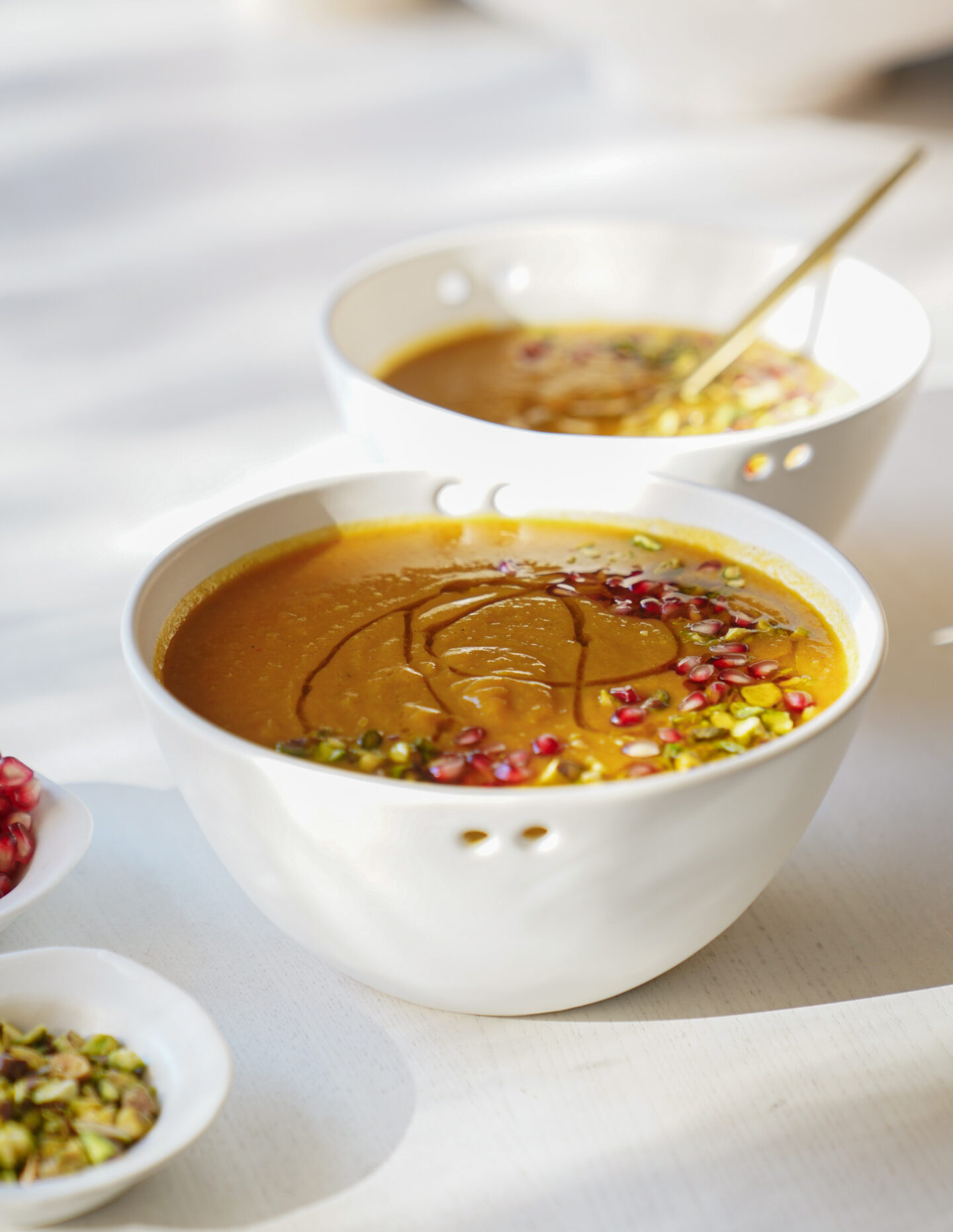 A white bowl of orange soup, topped with a drizzle of oil, pomegranate seeds, and chopped pistachios, sits on a light table with another similar bowl and small dishes in the background.