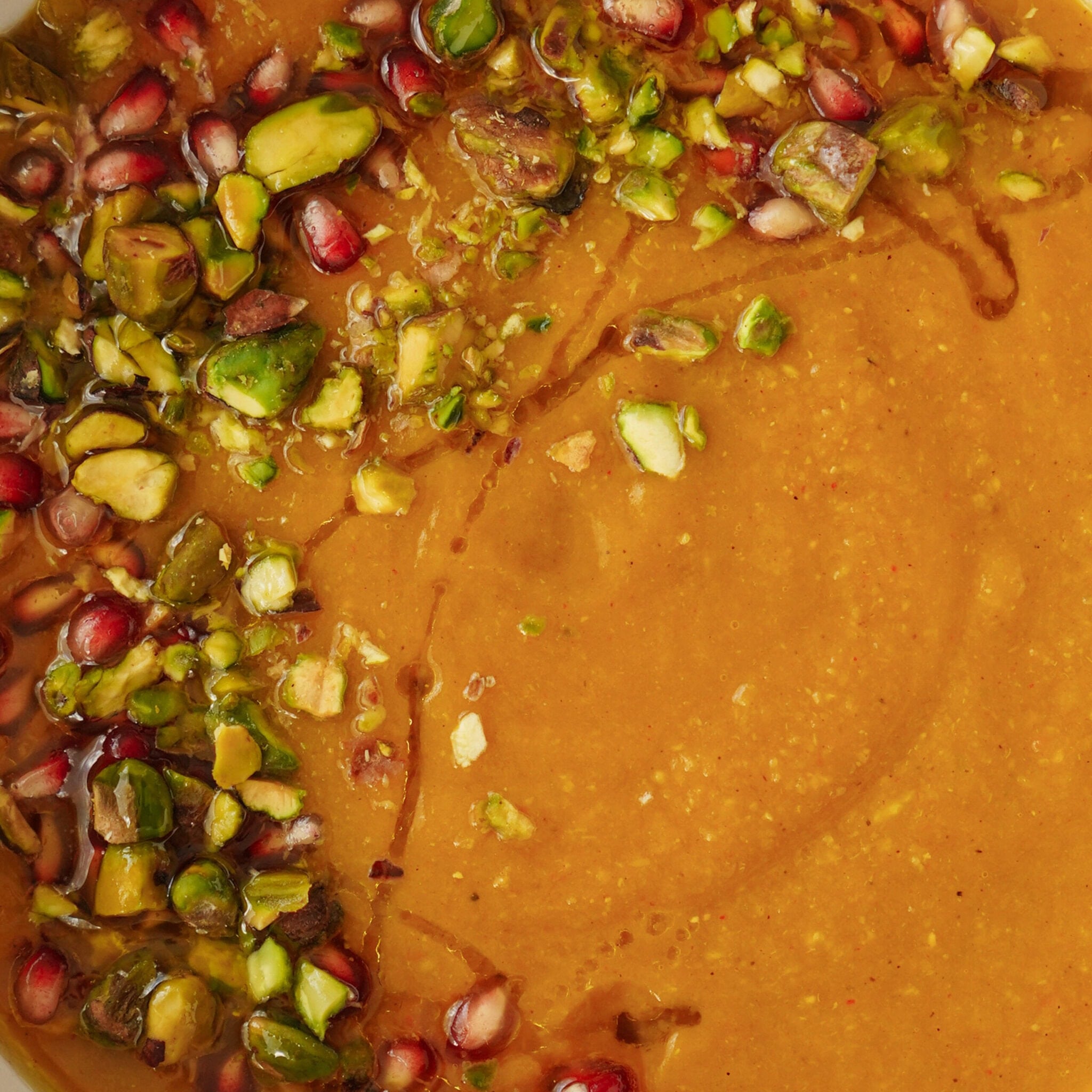 A close-up of a bowl of creamy soup garnished with chopped pistachios, pomegranate seeds, and a drizzle of oil. The vibrant toppings contrast with the smooth, orange-brown soup base.