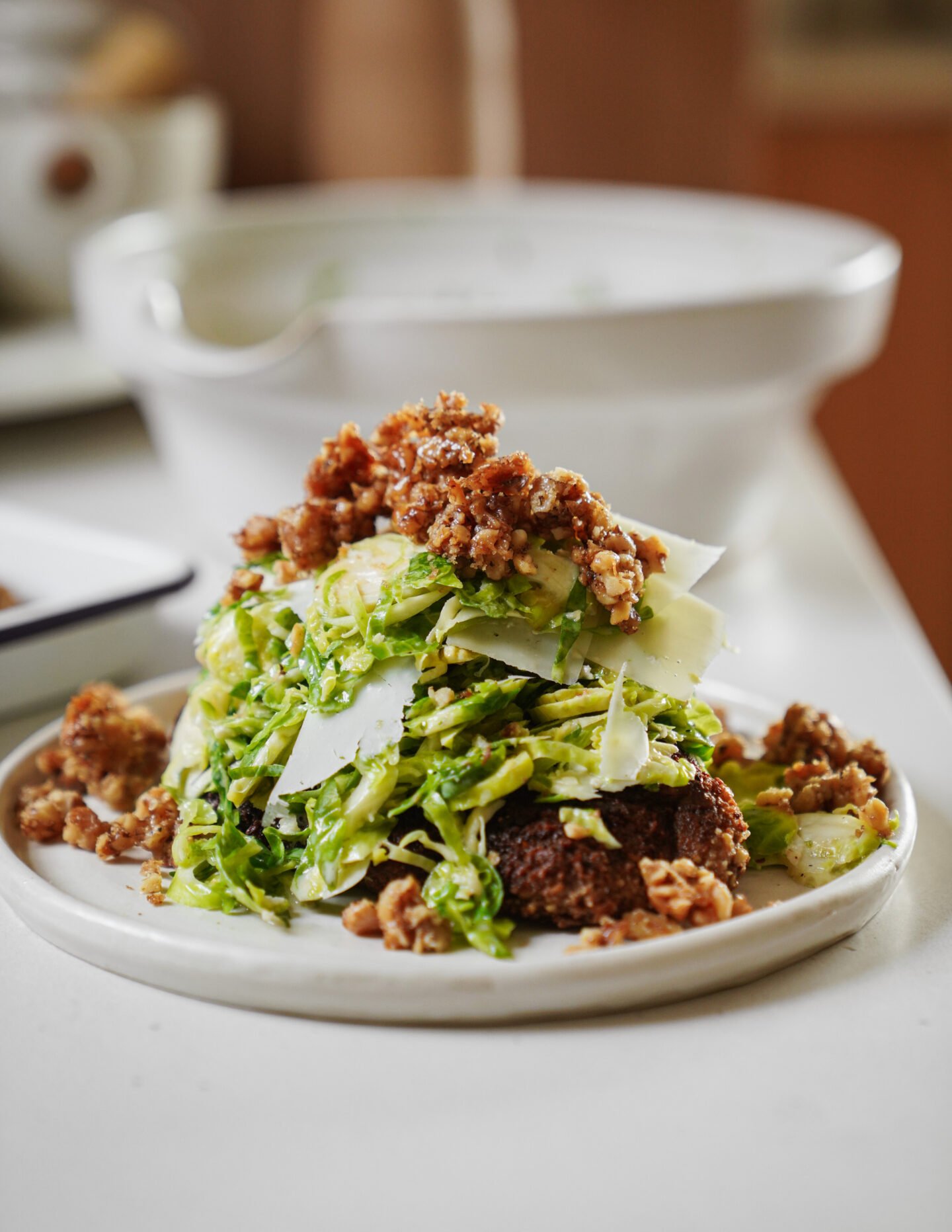 A plate of shaved Brussels sprouts salad topped with Parmesan shavings and crispy crumbled sausage, served on a slice of toasted bread. A white bowl is blurred in the background.