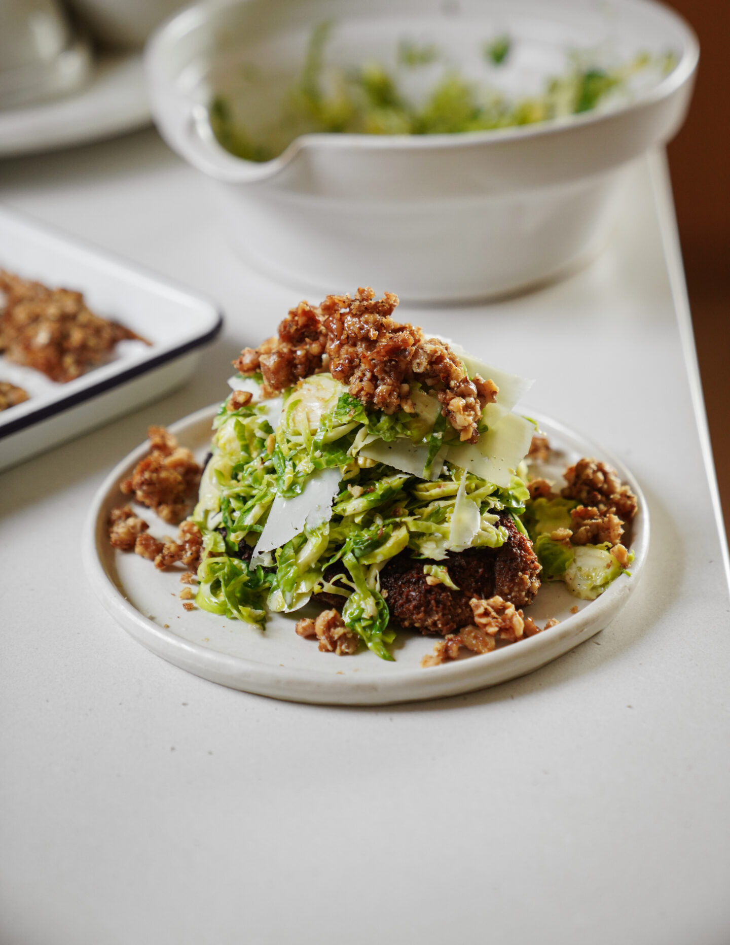 A plated dish featuring a slice of dark bread topped with shredded greens, shaved cheese, and crunchy caramelized nuts, served on a white plate with a white bowl and tray in the background.