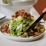 A close-up of a plate with a leafy green salad topped with shaved cheese and crunchy crumbled nuts, being cut with a fork and knife.