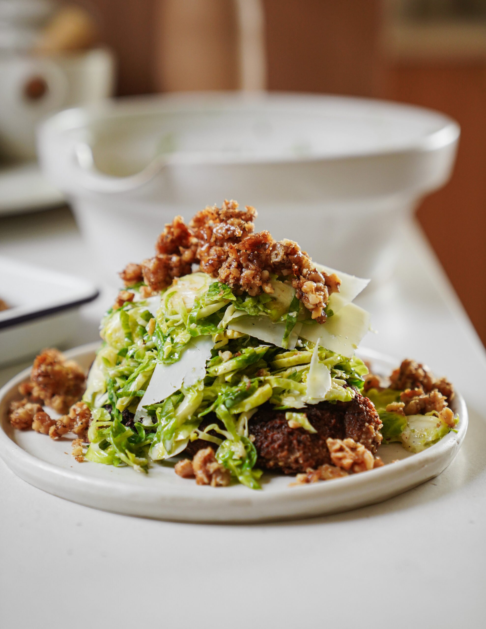 A plate of shaved Brussels sprouts salad topped with Parmesan shavings and crispy crumbled sausage, served on a slice of toasted bread. A white bowl is blurred in the background.