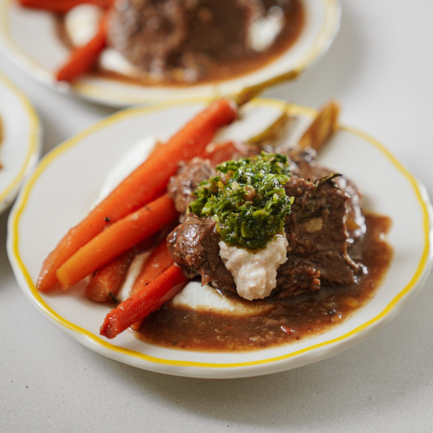 A plated dish featuring braised beef in brown sauce, topped with green herb garnish and a dollop of cream, served with glazed carrots on a white plate with a yellow rim.