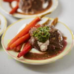 A plated dish featuring braised beef in brown sauce, topped with green herb garnish and a dollop of cream, served with glazed carrots on a white plate with a yellow rim.