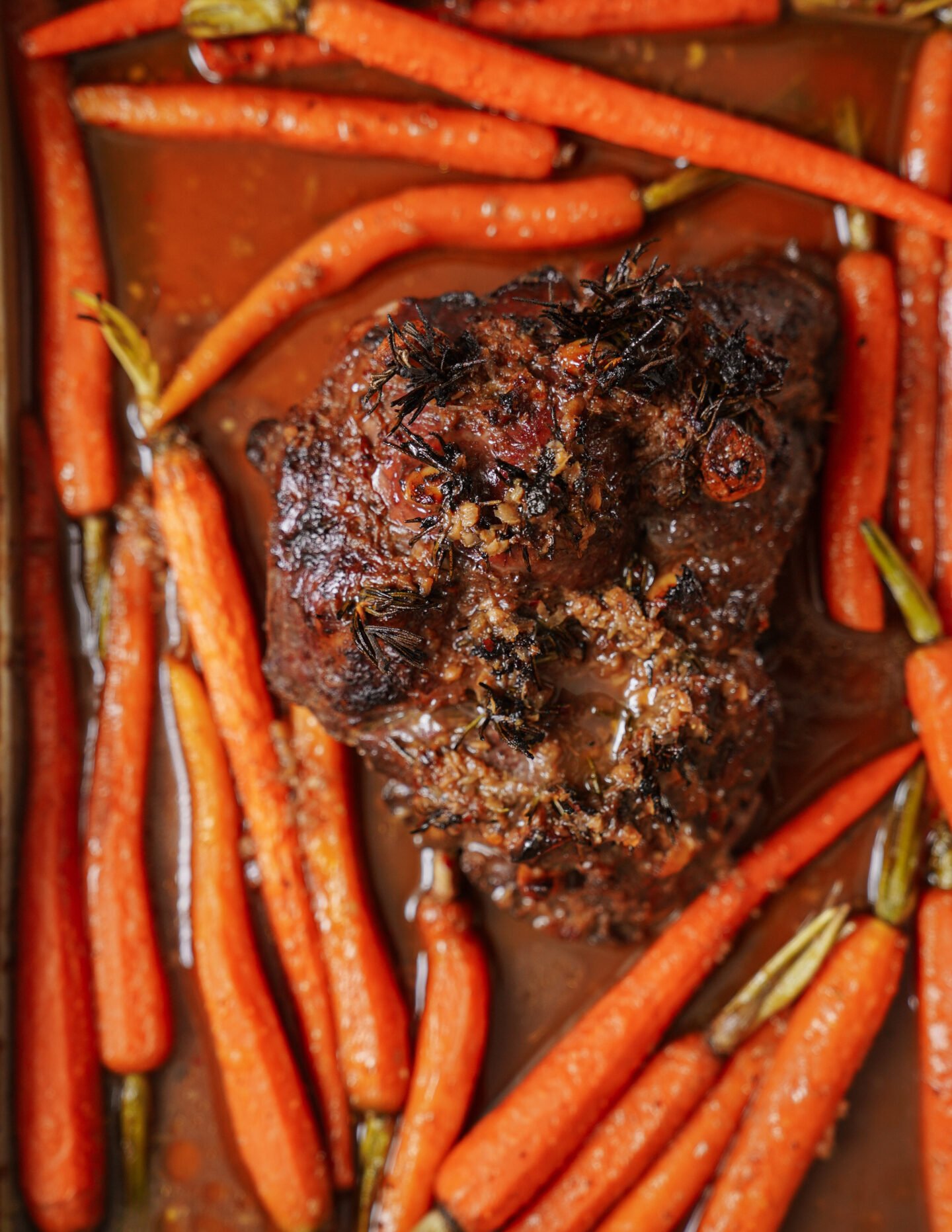 A roasted beef roast garnished with herbs sits on a tray surrounded by cooked baby carrots and a rich, brown pan sauce.