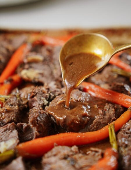A close-up of a golden spoon pouring gravy over a piece of cooked beef, surrounded by roasted carrots and garnished with herbs.