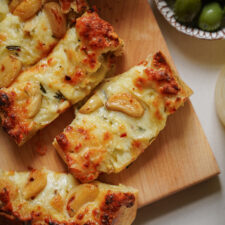 Close-up of sliced cheesy garlic bread topped with roasted garlic cloves on a wooden board, with a bowl of green olives partially visible in the background.