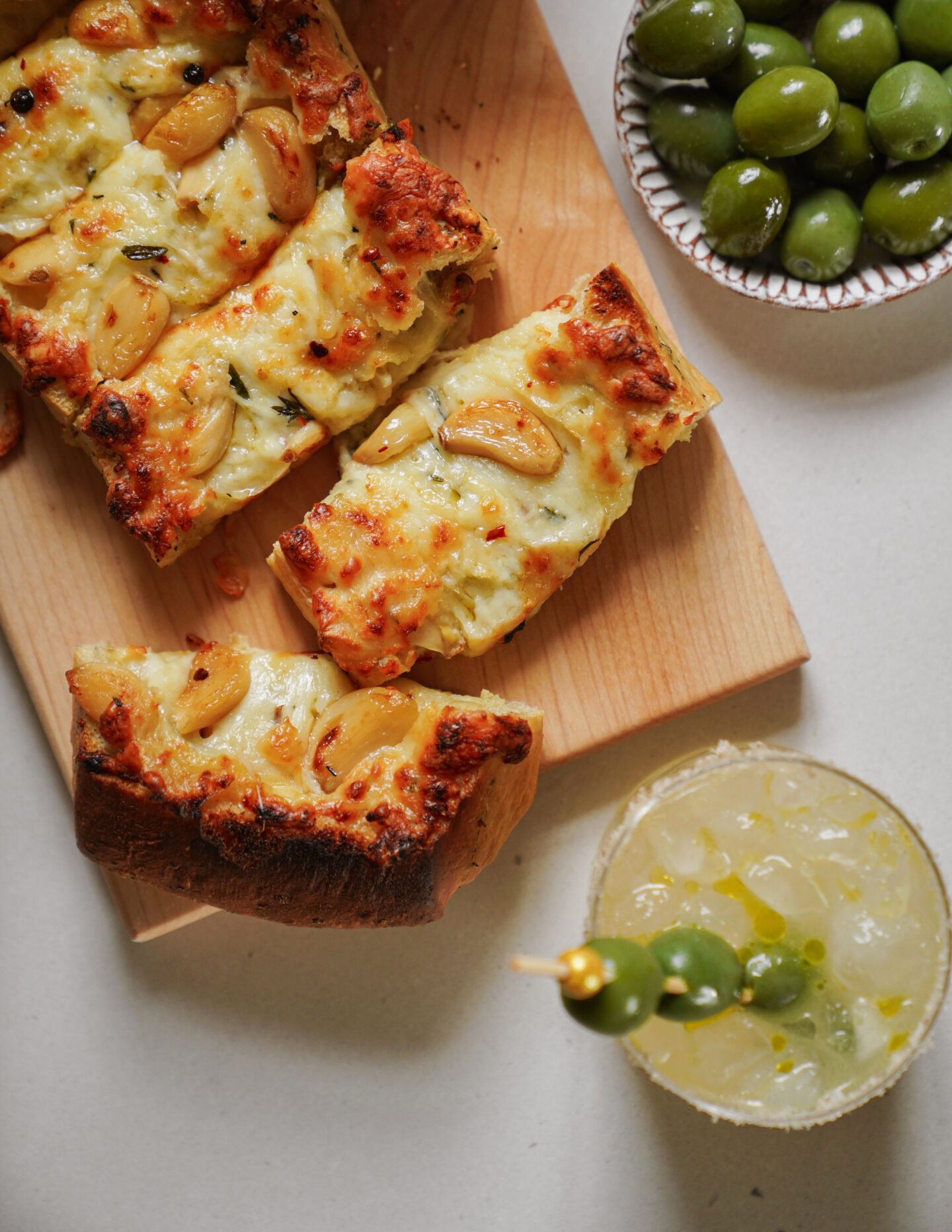 Three slices of cheesy garlic bread on a wooden board, a small bowl of green olives, and a cocktail garnished with olives and a twist of lemon are arranged on a light surface.