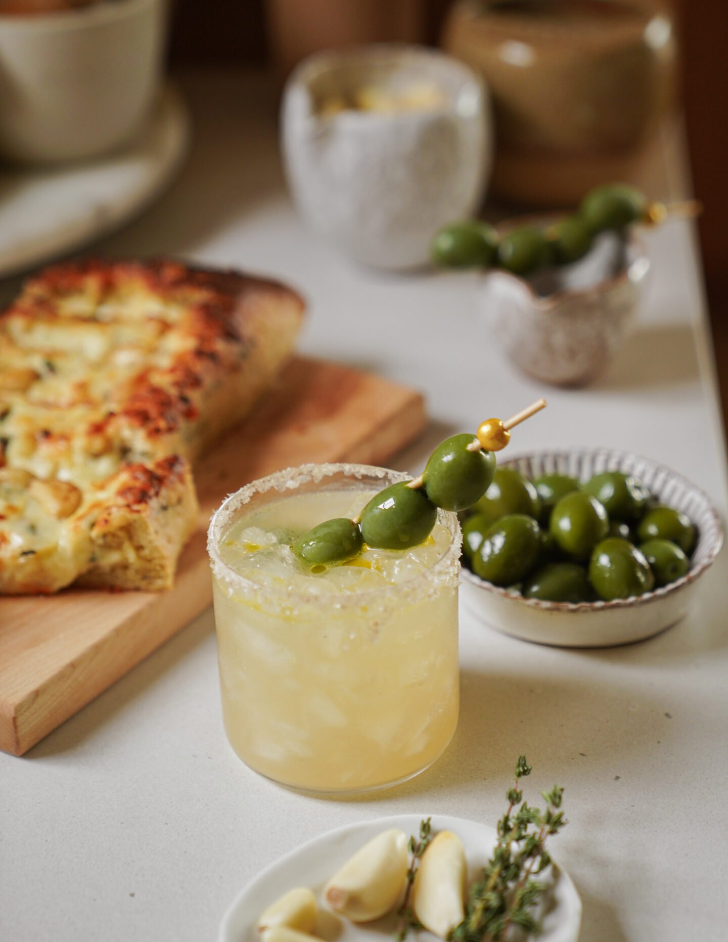 A cocktail garnished with green olives sits on a counter, next to a bowl of olives, garlic cloves, herbs, and a focaccia bread on a wooden board. Other small dishes are blurred in the background.
