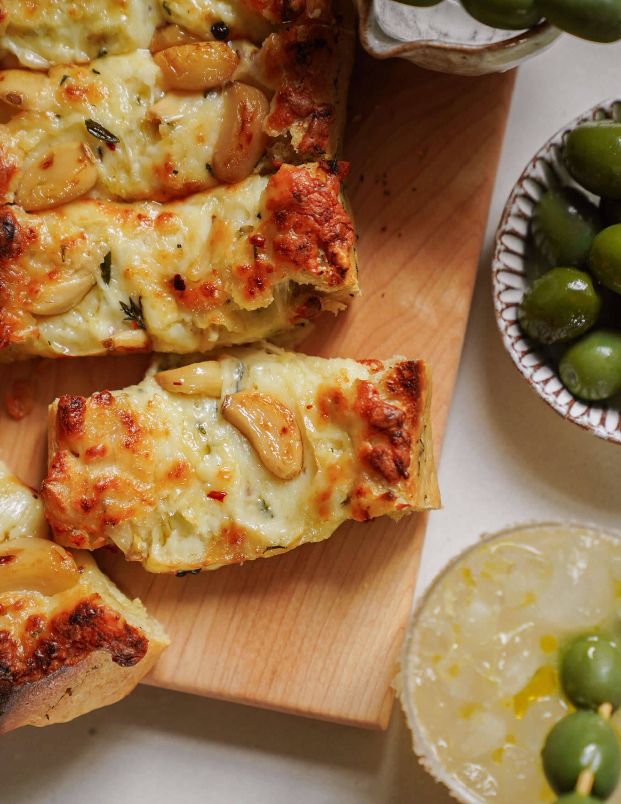 Slices of cheesy garlic bread topped with roasted garlic cloves on a wooden board, accompanied by bowls of green olives and a lemony-looking condiment.
