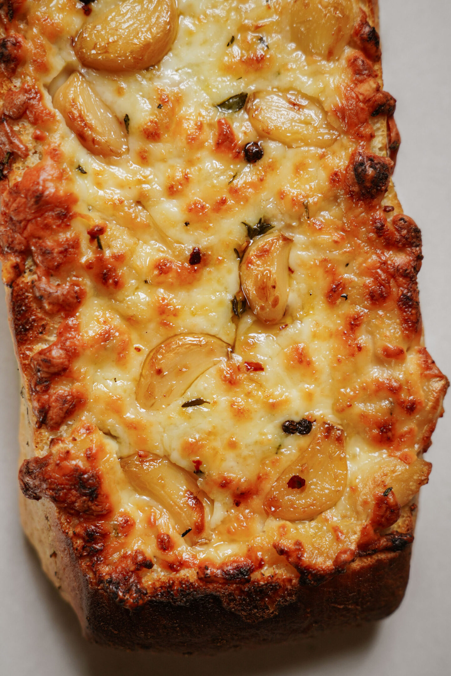 A close-up of a rectangular loaf of cheesy garlic bread, topped with melted cheese and whole roasted garlic cloves, with a golden-brown crust.