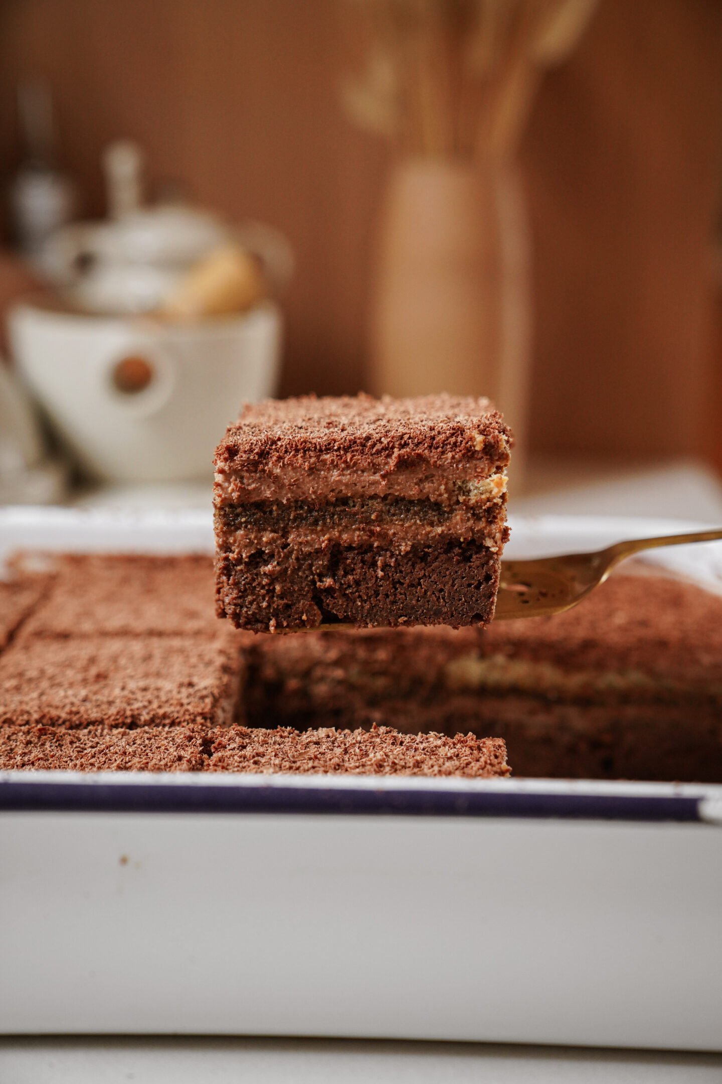 A close-up of a layered chocolate cake being lifted from a tray with a spatula, showing its distinct layers and a crumbly chocolate topping. A teapot and cups are blurred in the background.